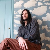 Man sitting against wall with long dark hair, wearing blue check shirt and brown trousers